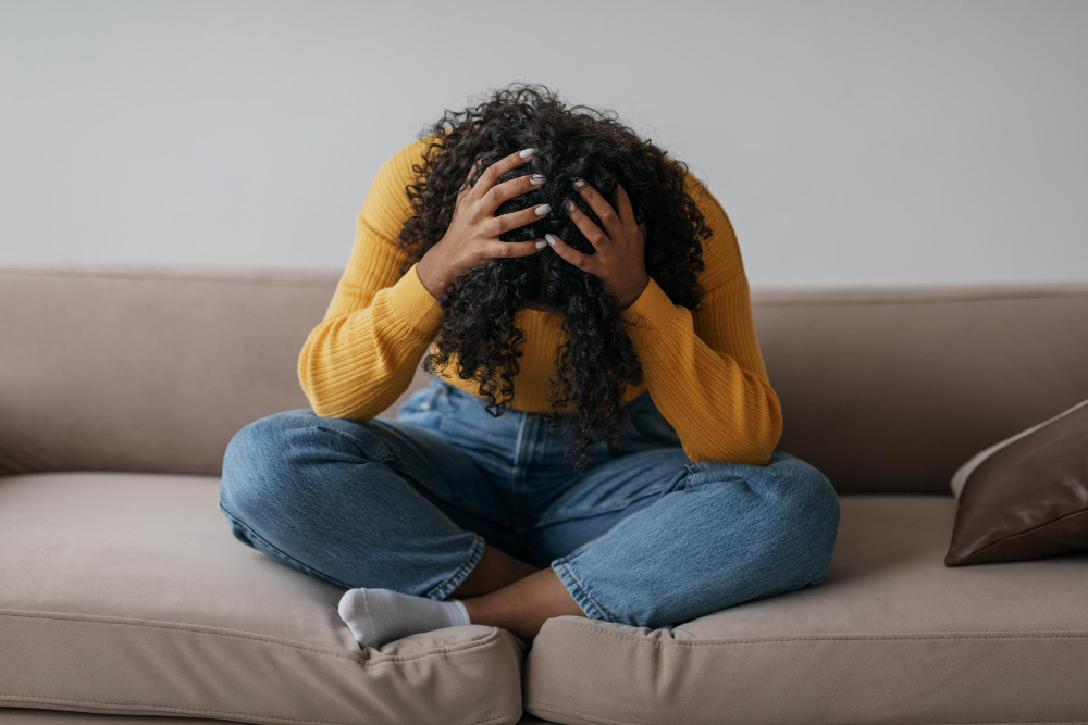 Depressed young black woman sitting cross legged on sofa with head in her hands at home
