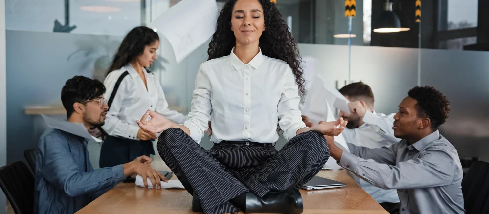 Calm businesswoman female leader woman sitting on table in lotus position meditating in office feels calmness ignore annoyed angry colleagues throw documents business conflict quarrel on background