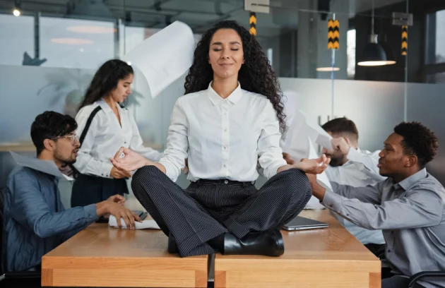 Calm businesswoman female leader woman sitting on table in lotus position meditating in office feels calmness ignore annoyed angry colleagues throw documents business conflict quarrel on background