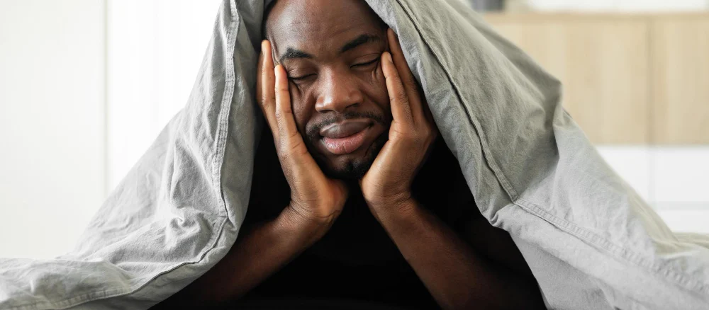 Sleepy African Guy Covered With Blanket Lying In Bedroom