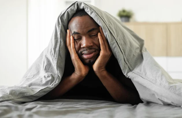 Sleepy African Guy Covered With Blanket Lying In Bedroom
