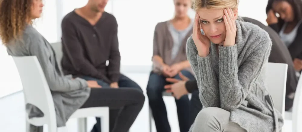 Upset woman with head in hands at rehab group looking at camera