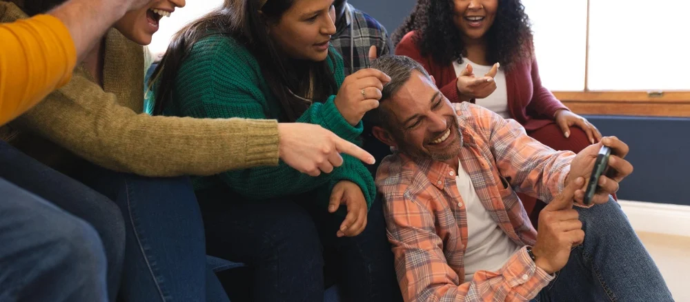 Happy diverse male and female friends relaxing at home together talking and looking at smartphone
