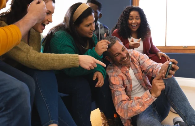 Happy diverse male and female friends relaxing at home together talking and looking at smartphone