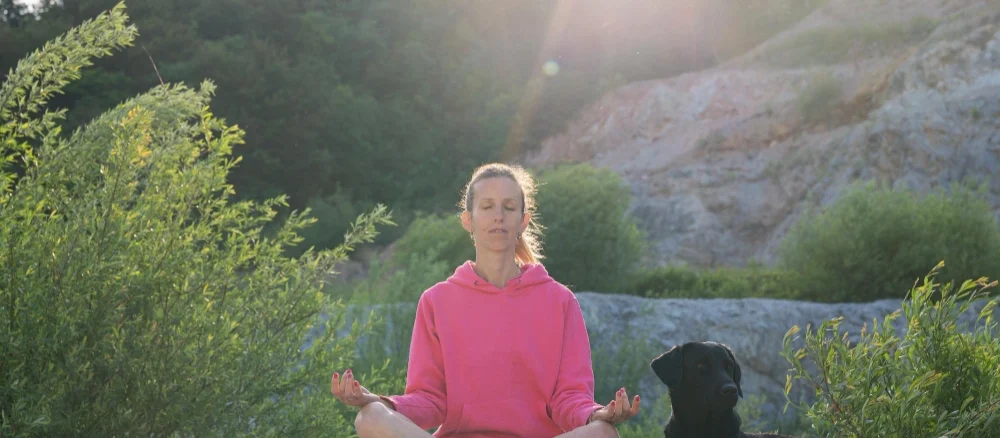 Young woman meditating in nature at sunrise with her dog beside her